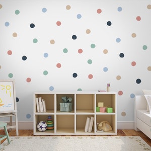 May include: A white wall with a colorful polka dot pattern. The wall is decorated with blue, green, pink, and brown dots. There is a white bookshelf with a few items on the shelves, a white easel with a drawing of a boat and sun, and a white bed with a light blue blanket.