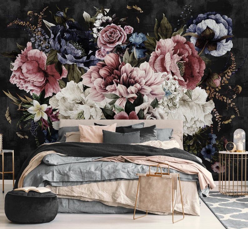 May include: A bedroom with a black and white floral wallpaper featuring large pink, white, and blue flowers. The bed has a black throw blanket, a light blue duvet, and pink and gray pillows. A gold metal side table with a glass cloche sits in the corner.
