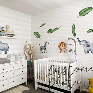 May include: A white dresser with black knobs, a white cot with a white mattress, and a yellow chair in a nursery decorated with animal wall decals. The decals include a rhino, a lion, an elephant, and a zebra. The decals are surrounded by green leaves.