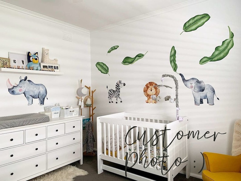 May include: A white dresser with black knobs, a white cot with a white mattress, and a yellow chair in a nursery decorated with animal wall decals. The decals include a rhino, a lion, an elephant, and a zebra. The decals are surrounded by green leaves.