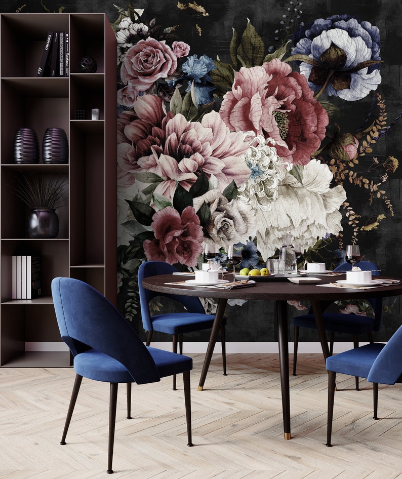 May include: A round wooden dining table with four blue velvet chairs. The table is set with plates, glasses, and silverware. The background is a dark floral wallpaper with large pink and white flowers.