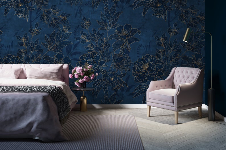 May include: A bedroom interior with a bed, a chair, and a floor lamp. The walls are covered in a blue wallpaper with a gold floral pattern. The bed has a pink headboard and a gray blanket. The chair is upholstered in pink fabric. The floor lamp has a gold base and a white shade.