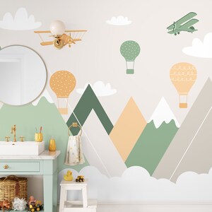 May include: A white wall with a green, orange, and gray mountain range design. The mountains are decorated with hot air balloons, a small airplane, and clouds. There is a mirror, a sink, and a towel rack in the foreground.