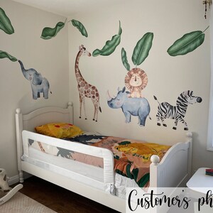 May include: A white toddler bed with a white bed rail and a colourful jungle animal themed bedding set. The wall behind the bed is decorated with jungle animal wall decals, including a giraffe, a lion, a rhino, a zebra, and an elephant. The decals are surrounded by green leaves.
