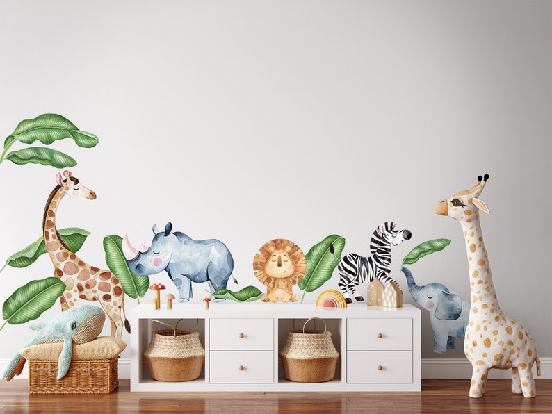 May include: A white wall with a set of jungle animal wall decals. The decals include a giraffe, rhino, lion, zebra, elephant, and banana leaves. The decals are on a white wall with a white dresser with three drawers and two baskets underneath.