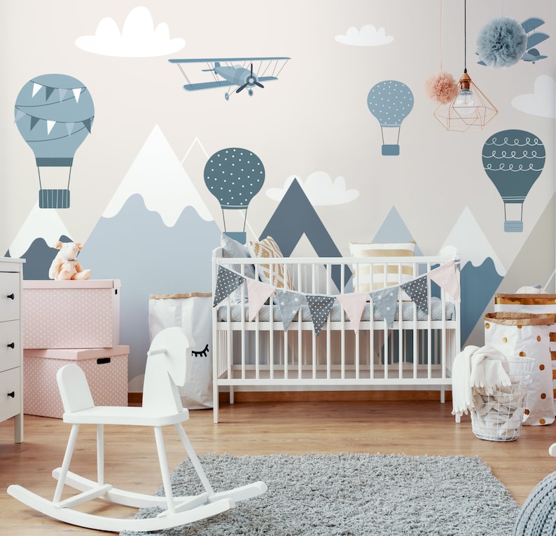 May include: A white crib with a blue and white patterned bunting hanging above it. The wall behind the crib is decorated with a mural of mountains, clouds, hot air balloons, and a plane. The room is decorated in a blue and white color scheme.