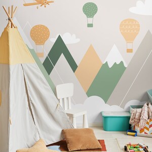 May include: A light gray teepee tent with a yellow top is set up in a playroom with a white chair and a blue storage bin. The wall is decorated with a mountain range mural with hot air balloons and airplanes.