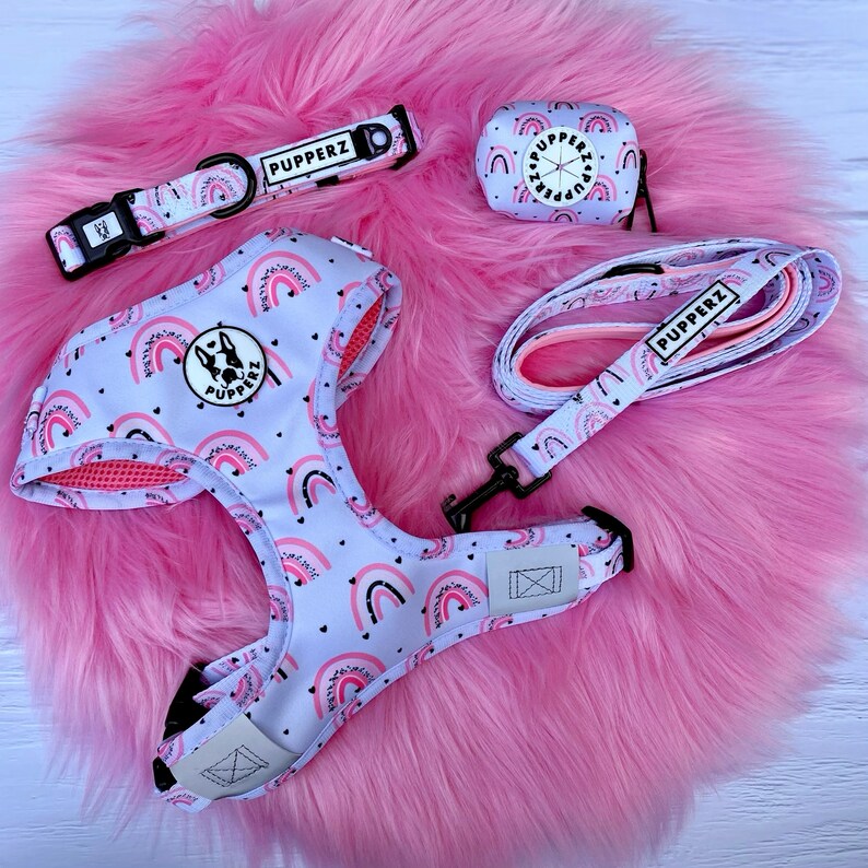 May include: A white dog harness with a rainbow and heart pattern, a matching leash, a pink and white poop bag holder, and a pink and white collar with the word "PUPPERZ" on it.