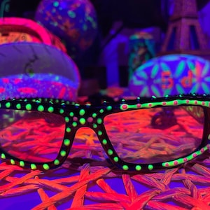 Op de afbeelding: Zwarte zonnebril met neon groene en roze stippen. De zonnebril ligt op een geweven oppervlak.