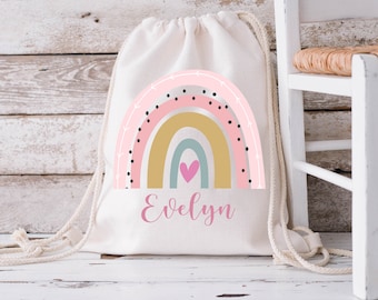 May include: A white drawstring bag with a pink, yellow, and blue rainbow design. The rainbow has black polka dots and white arrows. The bag has the name "Evelyn" printed in pink below the rainbow.