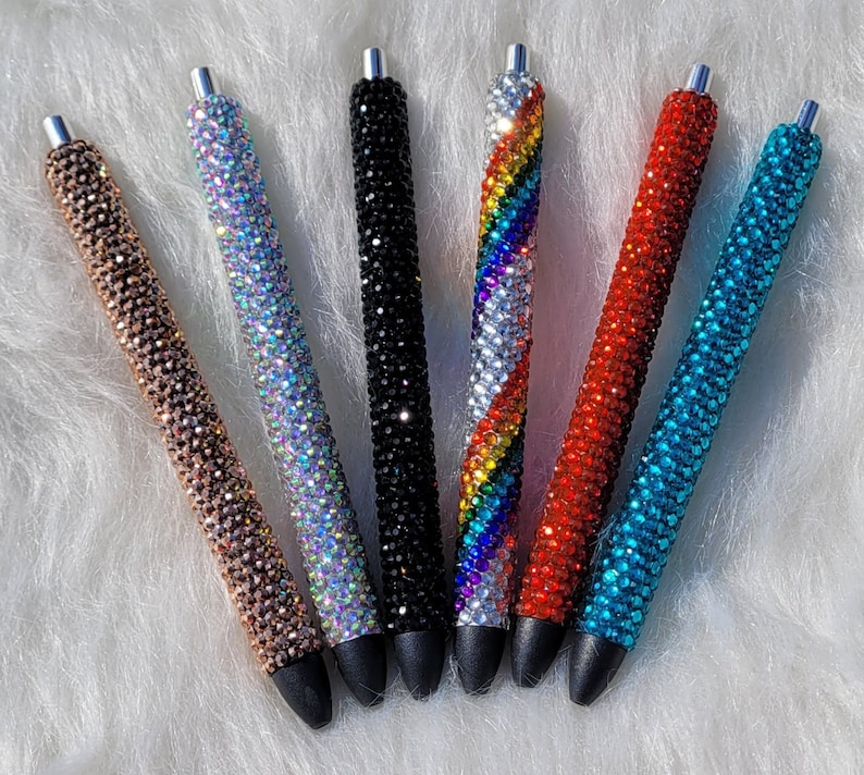 May include: Seven rhinestone pens in various colors, including black, red, blue, rainbow, and iridescent. The pens are all decorated with rhinestones.