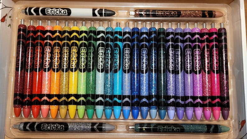 May include: A set of 30 glitter crayons in a variety of colors, including red, orange, yellow, green, blue, purple, and pink. The crayons are packaged in a clear plastic case with the brand name "Ericka" printed on the top and bottom.
