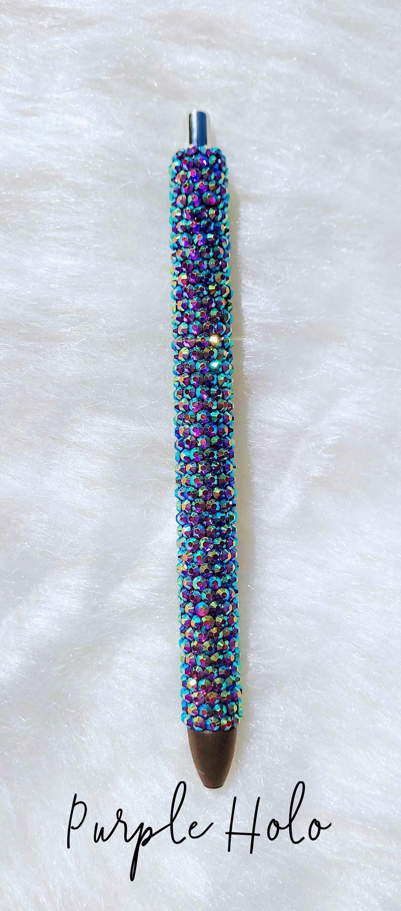 May include: A purple holographic pen covered in iridescent rhinestones. The pen is on a white fluffy surface. "Purple Holo" is written in black below the pen.