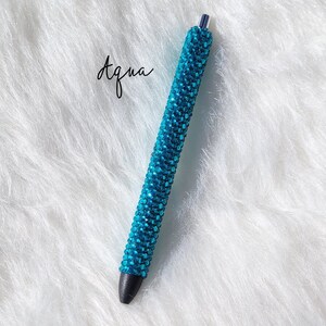 May include: A turquoise blue pen covered in rhinestones. The pen has a black tip and is on a white fluffy surface.