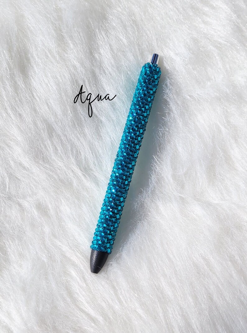 May include: A turquoise blue pen covered in rhinestones. The pen has a black tip and is on a white fluffy surface.