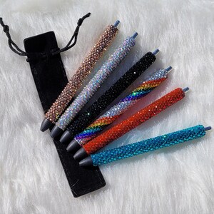 May include: Set of seven rhinestone pens in a black velvet pouch. The pens are in various colors, including turquoise, red, rainbow, black, iridescent, and rose gold.