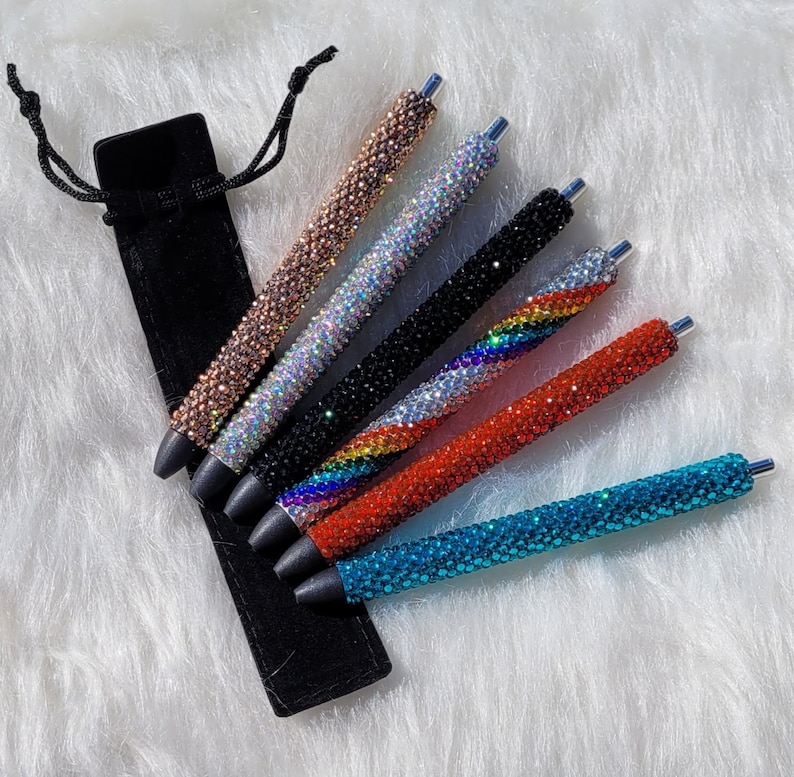 May include: Set of seven rhinestone pens in a black velvet pouch. The pens are in various colors, including turquoise, red, rainbow, black, iridescent, and rose gold.
