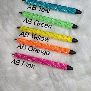 May include: Six rhinestone pens in different colors: purple, teal, green, yellow, orange, and pink.  The pens are arranged vertically, with the purple pen at the top and the pink pen at the bottom.  The pens are all the same size and shape, and they have a silver metal clip.