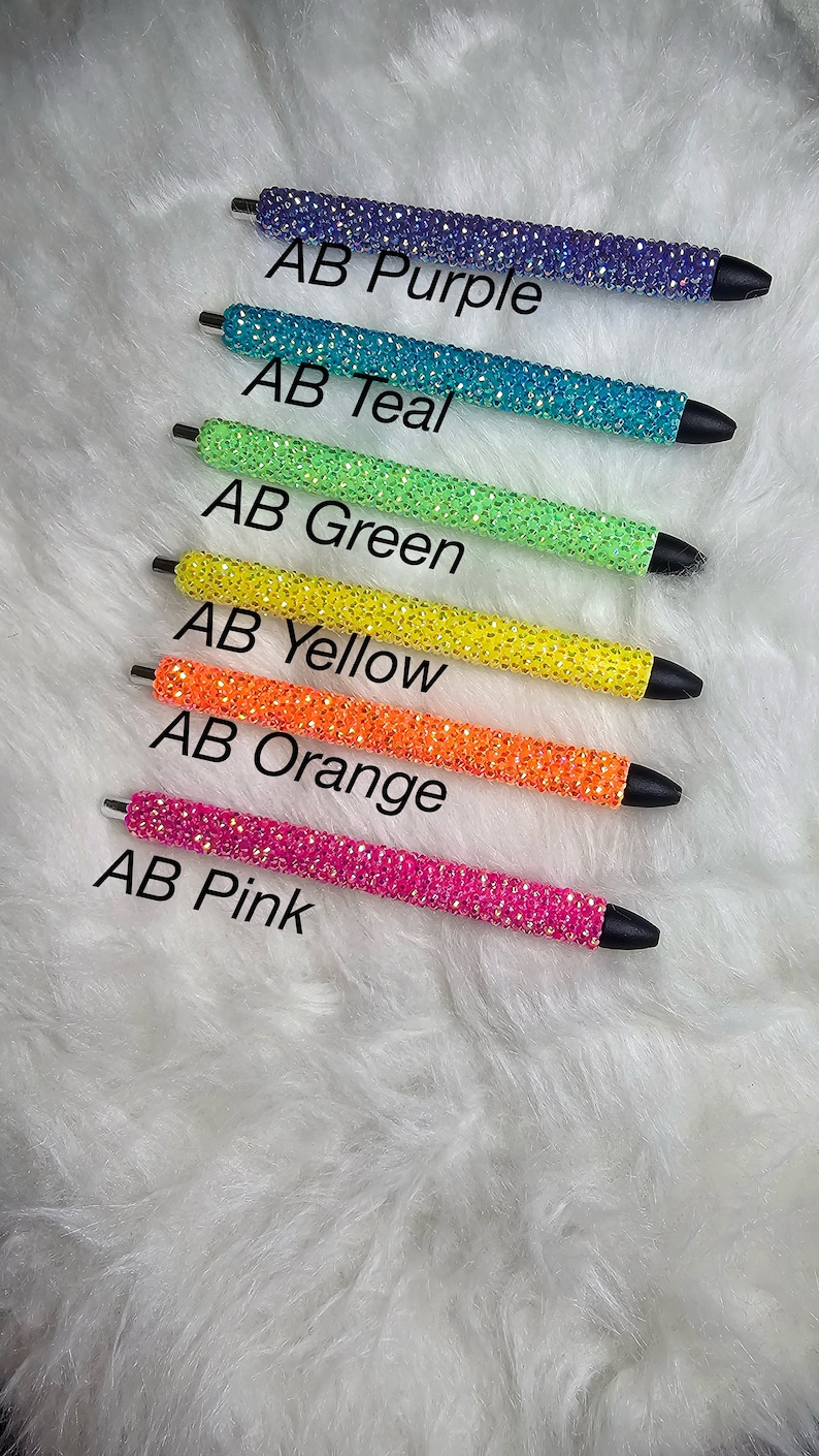 May include: Six rhinestone pens in different colors: purple, teal, green, yellow, orange, and pink.  The pens are arranged vertically, with the purple pen at the top and the pink pen at the bottom.  The pens are all the same size and shape, and they have a silver metal clip.