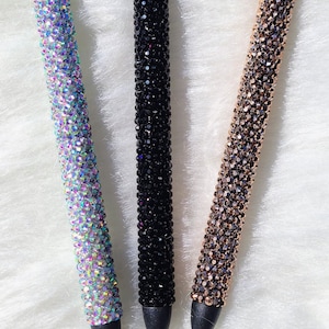 May include: Three rhinestone pens in different colors: silver holo, onyx, and rose gold. The pens are on a white fuzzy surface.