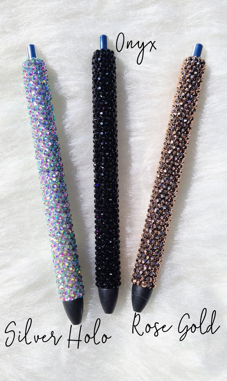 May include: Three rhinestone pens in different colors: silver holo, onyx, and rose gold. The pens are on a white fuzzy surface.