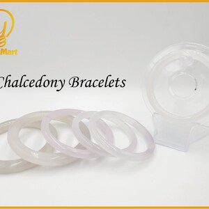 May include: Four white chalcedony bracelets of varying sizes, stacked on a white surface. The bracelets are smooth and polished, with a slight pink hue. The word "Chalcedony Bracelets" is written in black text above the bracelets.