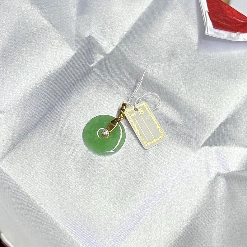 K&ouml;nnte beinhalten: Ein runder gr&uuml;ner Jadeanh&auml;nger mit einer goldfarbenen &Ouml;se und einem kleinen, klaren, rund geschliffenen Stein. Der Anh&auml;nger ist an einer wei&szlig;en Schnur mit einem rechteckigen wei&szlig;en Etikett befestigt, das einen goldenen Rand und den Text NO und Y hat.