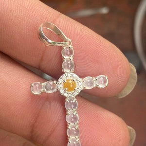 May include: A silver cross pendant with a central yellow gemstone surrounded by a halo of small white gemstones. The arms of the cross are adorned with additional white gemstones. The pendant is held by a silver loop.