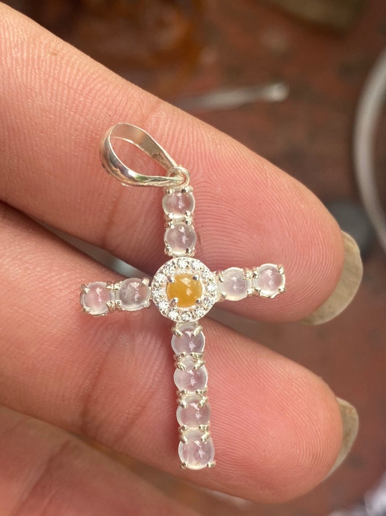 May include: A silver cross pendant with a central yellow gemstone surrounded by a halo of small white gemstones. The arms of the cross are adorned with additional white gemstones. The pendant is held by a silver loop.