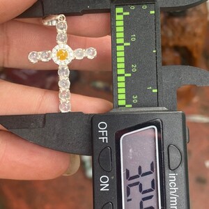 May include: A silver cross pendant with a central yellow gemstone and clear, round accents. The pendant is being measured with a caliper, showing a measurement of 32.4 mm. The cross is a religious symbol.