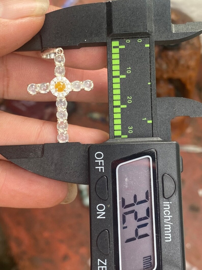 May include: A silver cross pendant with a central yellow gemstone and clear, round accents. The pendant is being measured with a caliper, showing a measurement of 32.4 mm. The cross is a religious symbol.