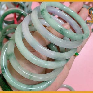 May include: A selection of jade bangles in shades of green and white. The smooth, polished bracelets are held in a hand, highlighting their translucent appearance. The bangles are a classic piece of jewellery.