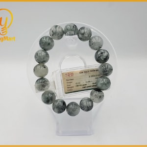 Op de afbeelding: Een grijze en witte edelsteen armband met ronde kralen. De armband is op een witte displaystandaard. Het label op de standaard luidt "CẢM THẠCH THIÊN N 5.3320.0125".