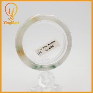 May include: A natural jadeite bangle bracelet. The bracelet is a light green color with areas of white and orange. A tag reads "NATURAL JADEITE" and "No: 43250". The bracelet is displayed on a clear stand.