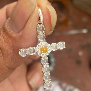 May include: A silver cross pendant featuring a central yellow gemstone surrounded by a halo of small clear stones. The cross arms are adorned with a series of clear, round stones. The pendant is held by a silver bail.