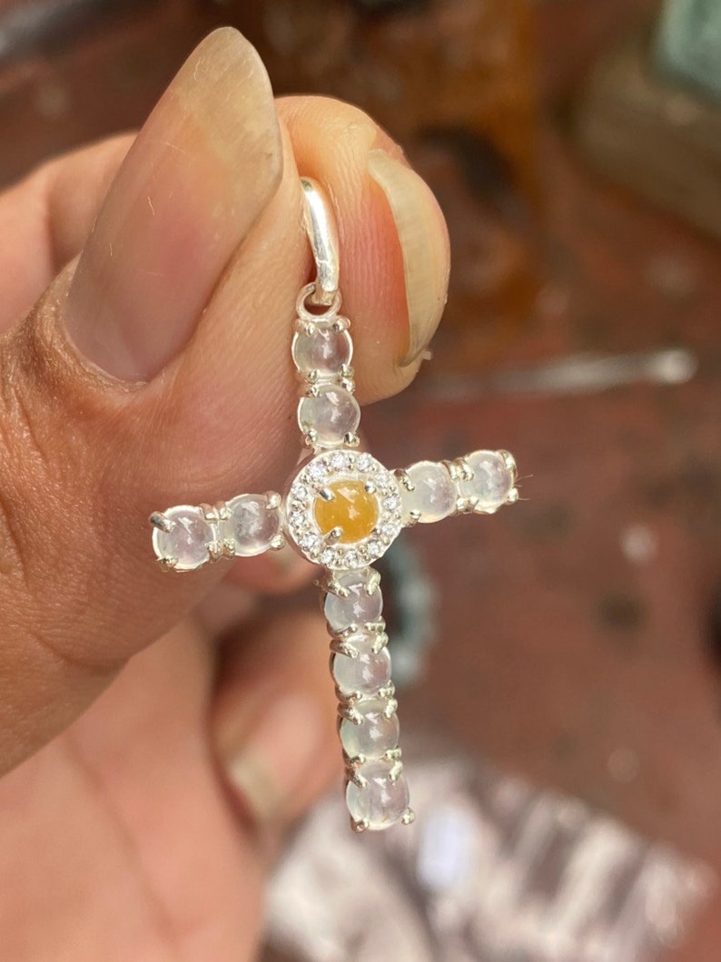 May include: A silver cross pendant featuring a central yellow gemstone surrounded by a halo of small clear stones. The cross arms are adorned with a series of clear, round stones. The pendant is held by a silver bail.