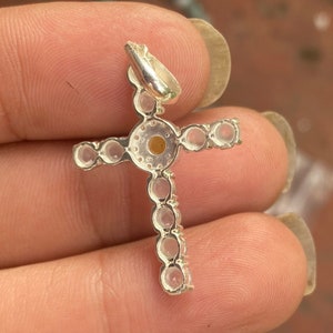 May include: A silver-colored cross pendant with a loop for a chain. The cross is adorned with small, round, clear gemstones. The center of the cross features a larger, round, amber-colored stone. The pendant is held in a hand.