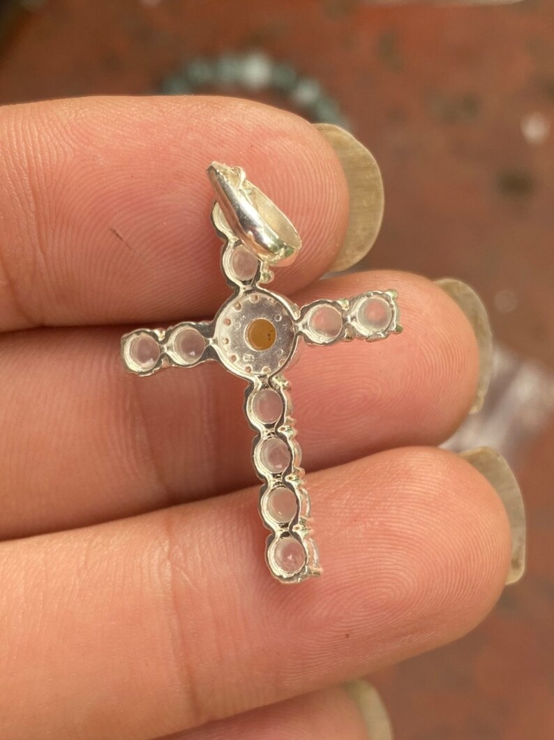 May include: A silver-colored cross pendant with a loop for a chain. The cross is adorned with small, round, clear gemstones. The center of the cross features a larger, round, amber-colored stone. The pendant is held in a hand.
