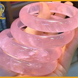 May include: A stack of five translucent, rose-colored quartz bangles. The smooth, polished bracelets are held in a hand, showcasing their soft pink hue and natural stone texture. The YingMart logo is visible in the upper left corner.