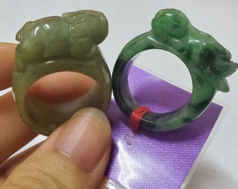 May include: Two jade rings featuring green and white jade stone. The rings are carved with animal designs. One ring has a green jade frog design and the other ring has a white jade animal design.