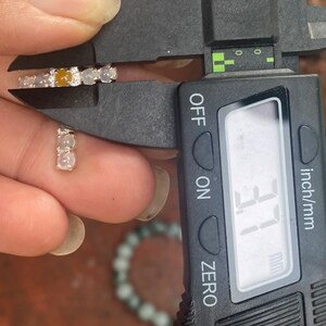 May include: A pair of small, silver earrings with a series of light-colored stones, including a central amber-colored stone. The earrings are being measured with a digital caliper, showing a measurement of 3.1 mm.