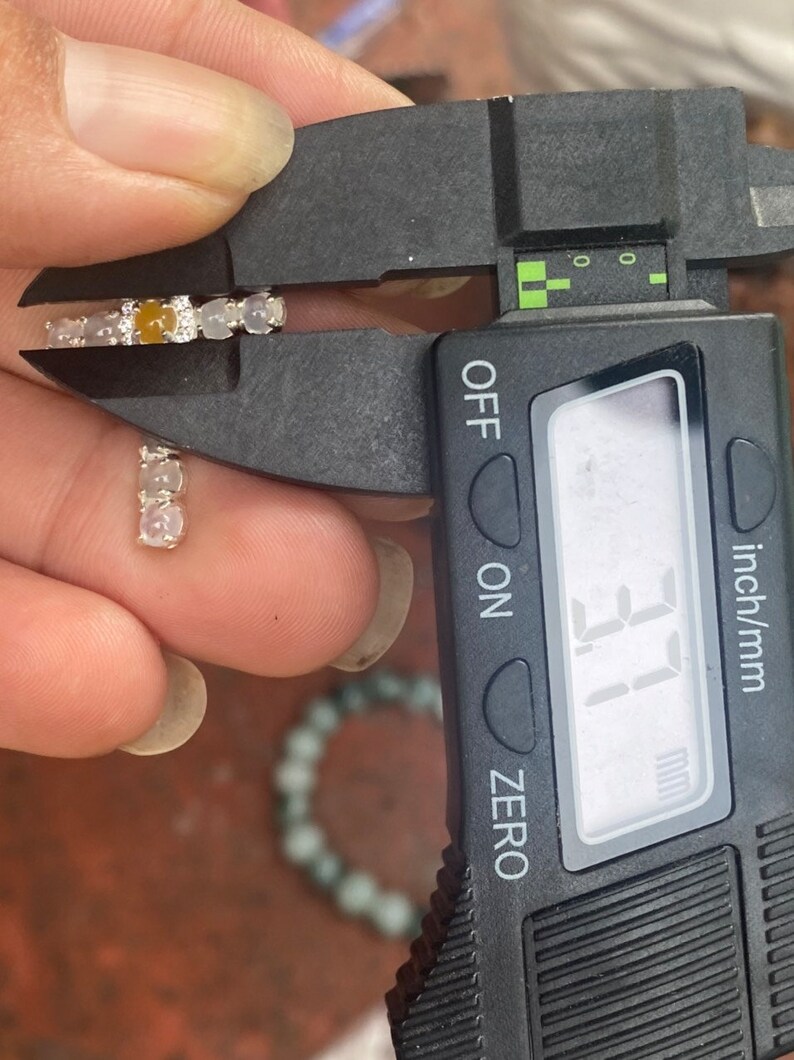 May include: A pair of small, silver earrings with a series of light-colored stones, including a central amber-colored stone. The earrings are being measured with a digital caliper, showing a measurement of 3.1 mm.