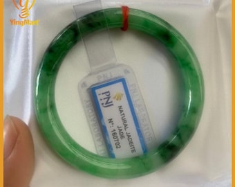 May include: A vibrant green jade bangle bracelet with dark green inclusions. The bracelet is displayed with a tag that reads "NATURAL JADEITE JADE" and "PNJ LABORATORY". The bracelet is a circular shape.