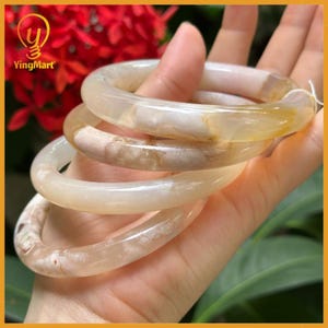 May include: Four translucent, light-colored stone bangles are stacked in a hand. The bangles have a mix of creamy white and light brown tones, with a smooth, polished finish. The background includes red flowers and the YingMart logo.