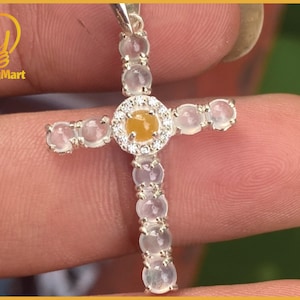May include: A silver cross pendant with a central yellow gemstone surrounded by small clear stones. The cross arms and vertical stem are adorned with clear, oval-shaped stones. The pendant is held by a person's fingers.