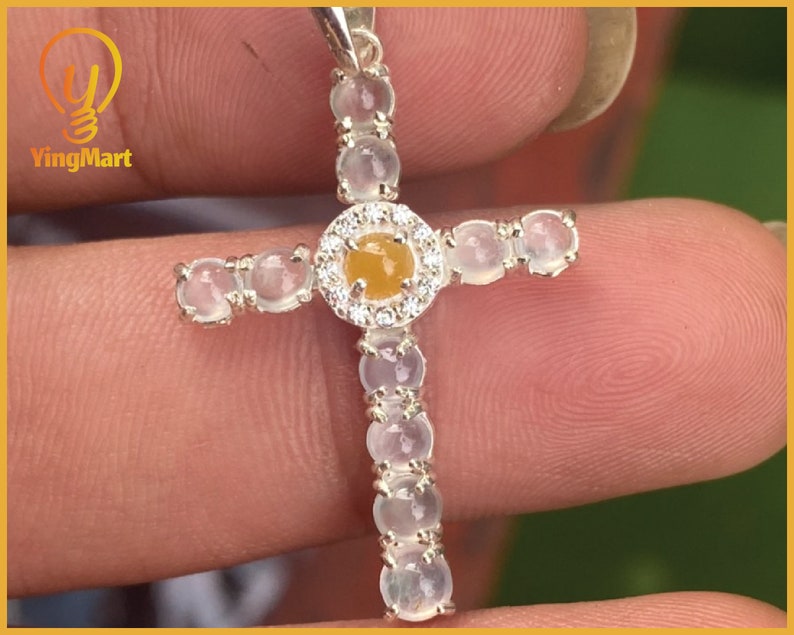 May include: A silver cross pendant with a central yellow gemstone surrounded by small clear stones. The cross arms and vertical stem are adorned with clear, oval-shaped stones. The pendant is held by a person's fingers.