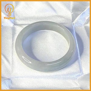 May include: A smooth, polished bangle bracelet crafted from translucent, pale greyish-white jadeite stone. The circular jewelry piece features a rounded profile with a glossy, reflective surface that catches light, showcasing a solid, seamless construction.
