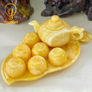 May include: A yellow marble tea set with a teapot and six cups, all sitting on a leaf-shaped tray. The set is made of a translucent yellow marble with white veins.