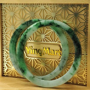 May include: Two jade bangles with green and white marbling are displayed in a gold-colored box. The box has a geometric pattern and the text "YingMart" in gold lettering. The bangles are circular and appear to be made of polished stone.