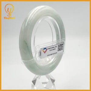 May include: A light green jade bangle bracelet displayed on a clear stand. The bracelet has a smooth, polished surface with subtle color variations. A tag with the text "NATURAL FEI CUI JADE" is attached to the stand.